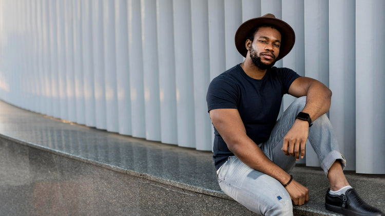 Man sitting against a modern architectural wall wearing a black shirt, light jeans, and a brown hat.