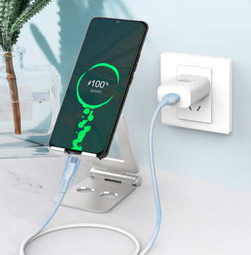 Phone charging on a stand with a power adapter plugged into a wall socket, on a light blue gradient background.