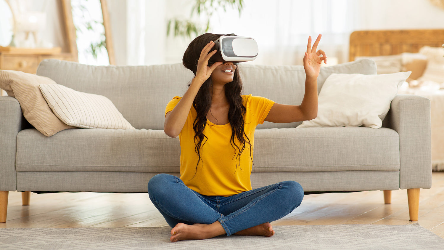 Woman wearing a yellow shirt and blue jeans sitting on the floor using a VR headset in a living room.